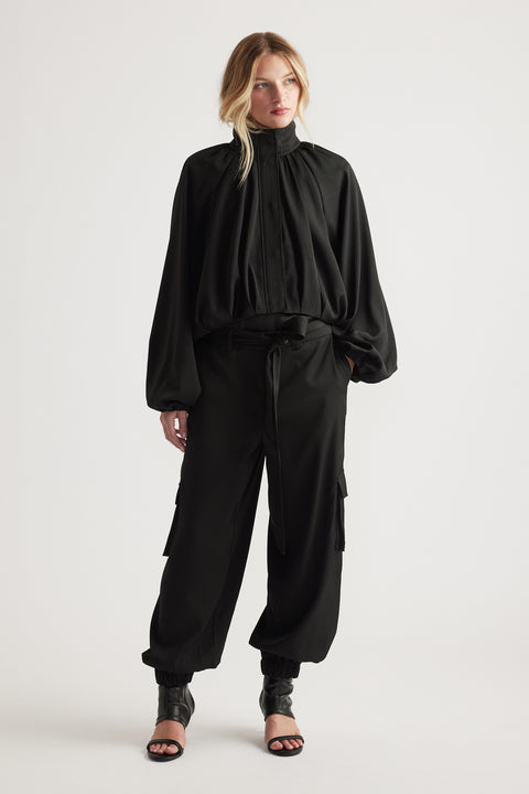 Woman wearing flowing black blouse with high neckline paired with relaxed fit black satin cargo pants and heels
