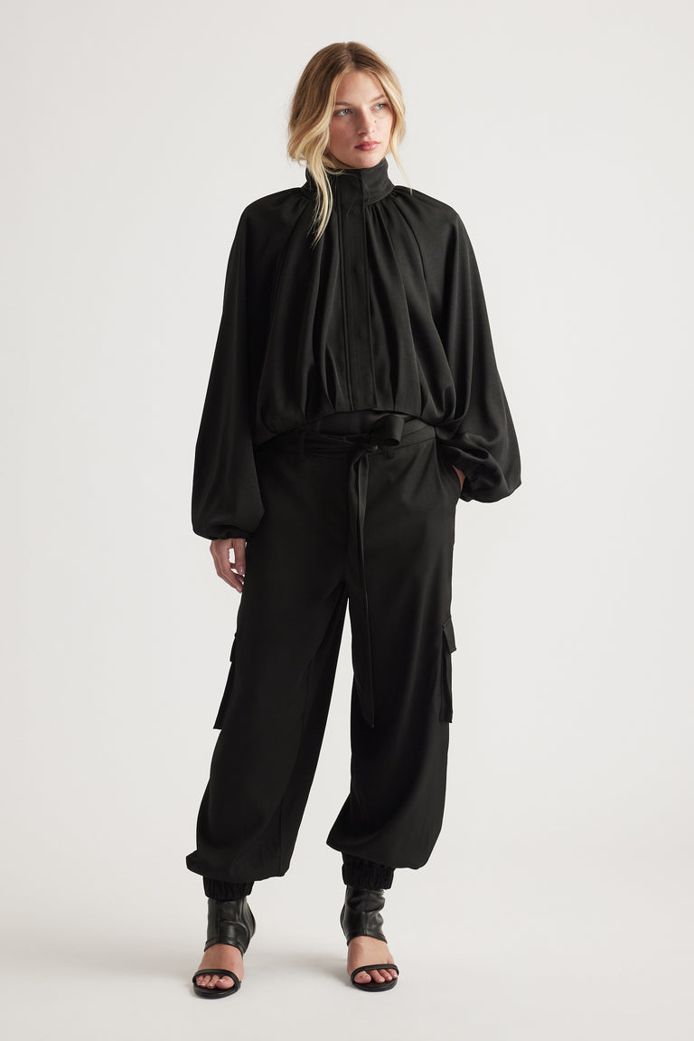 Woman wearing flowing black blouse with high neckline paired with relaxed fit black satin cargo pants and heels