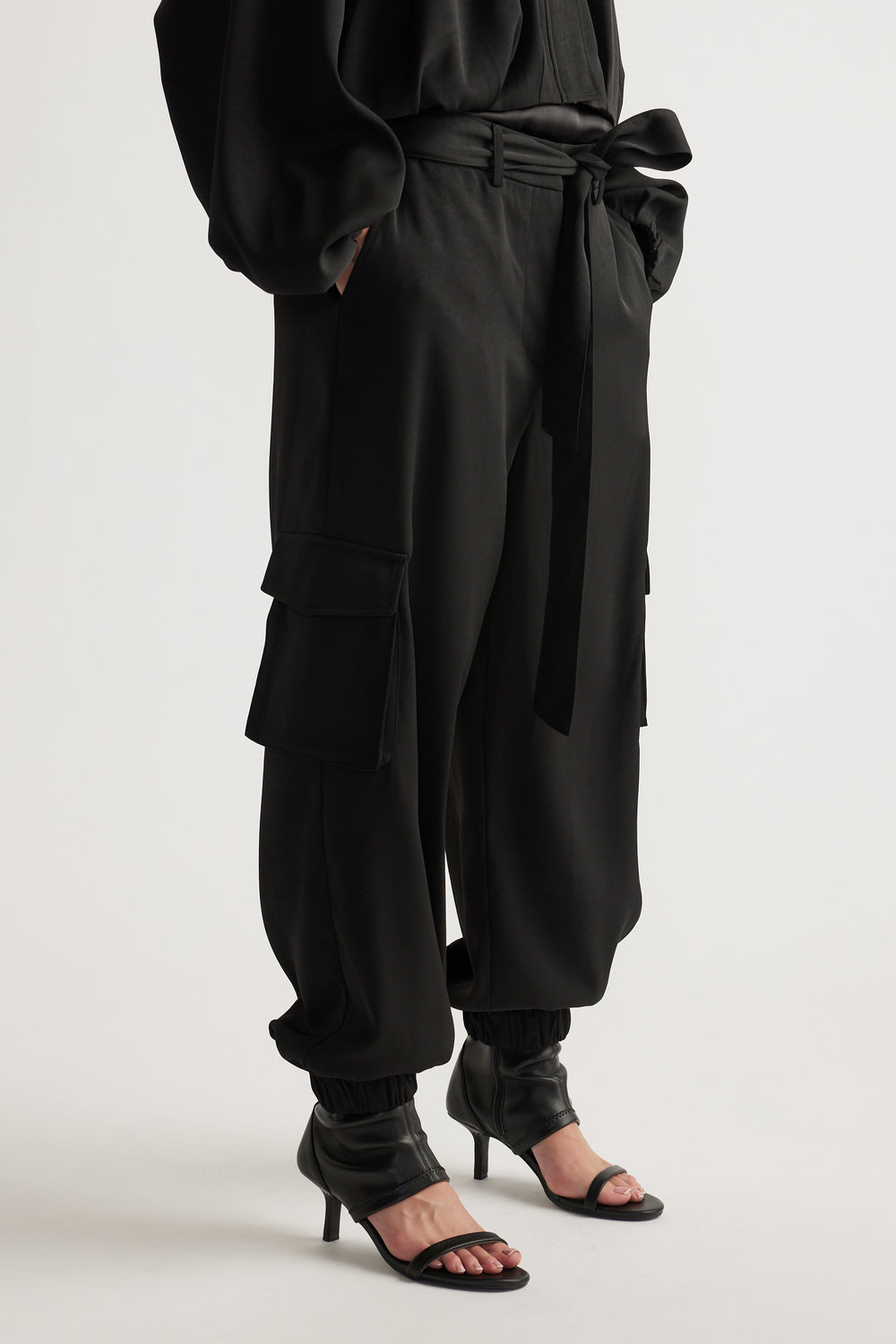 Black satin cargo pants with relaxed fit, tie waist, cuffed ankles, and side flap pockets styled with open-toe heels