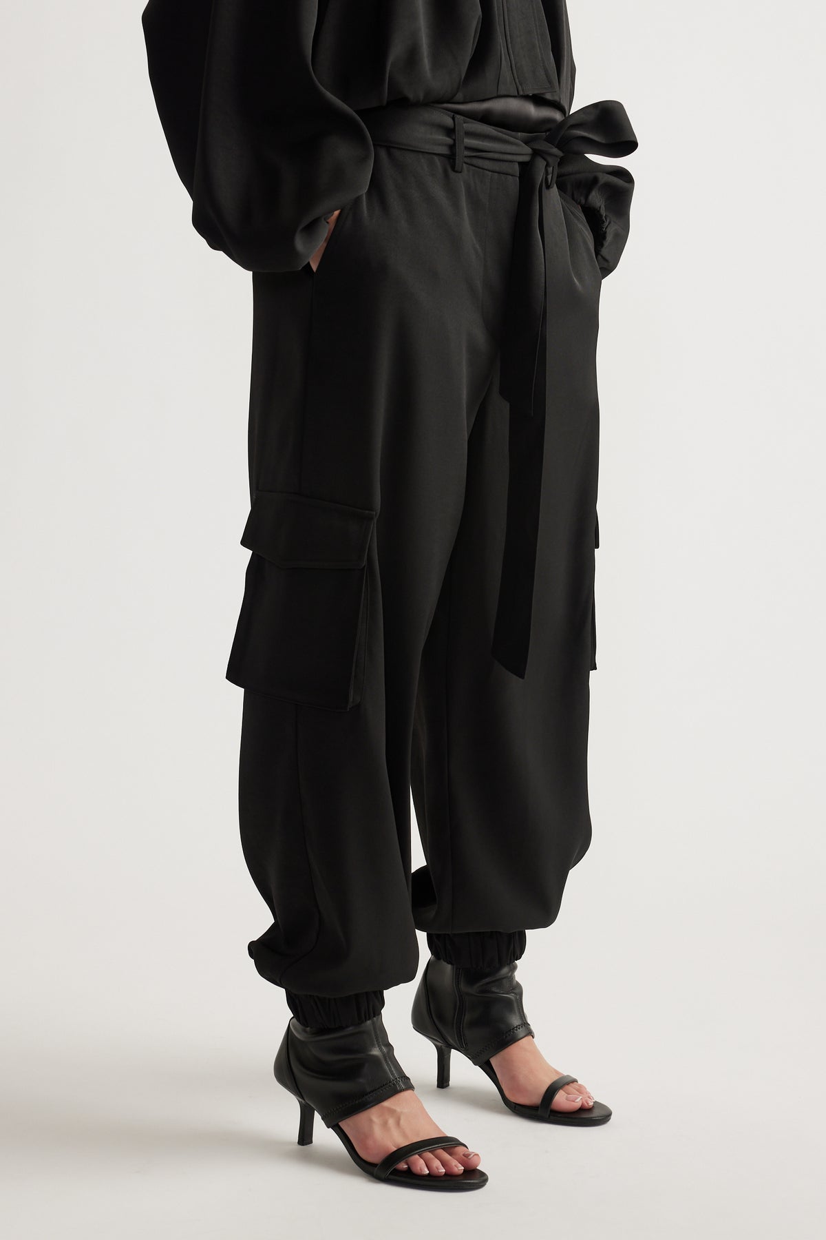 Black satin cargo pants with relaxed fit, tie waist, cuffed ankles, and side flap pockets styled with open-toe heels