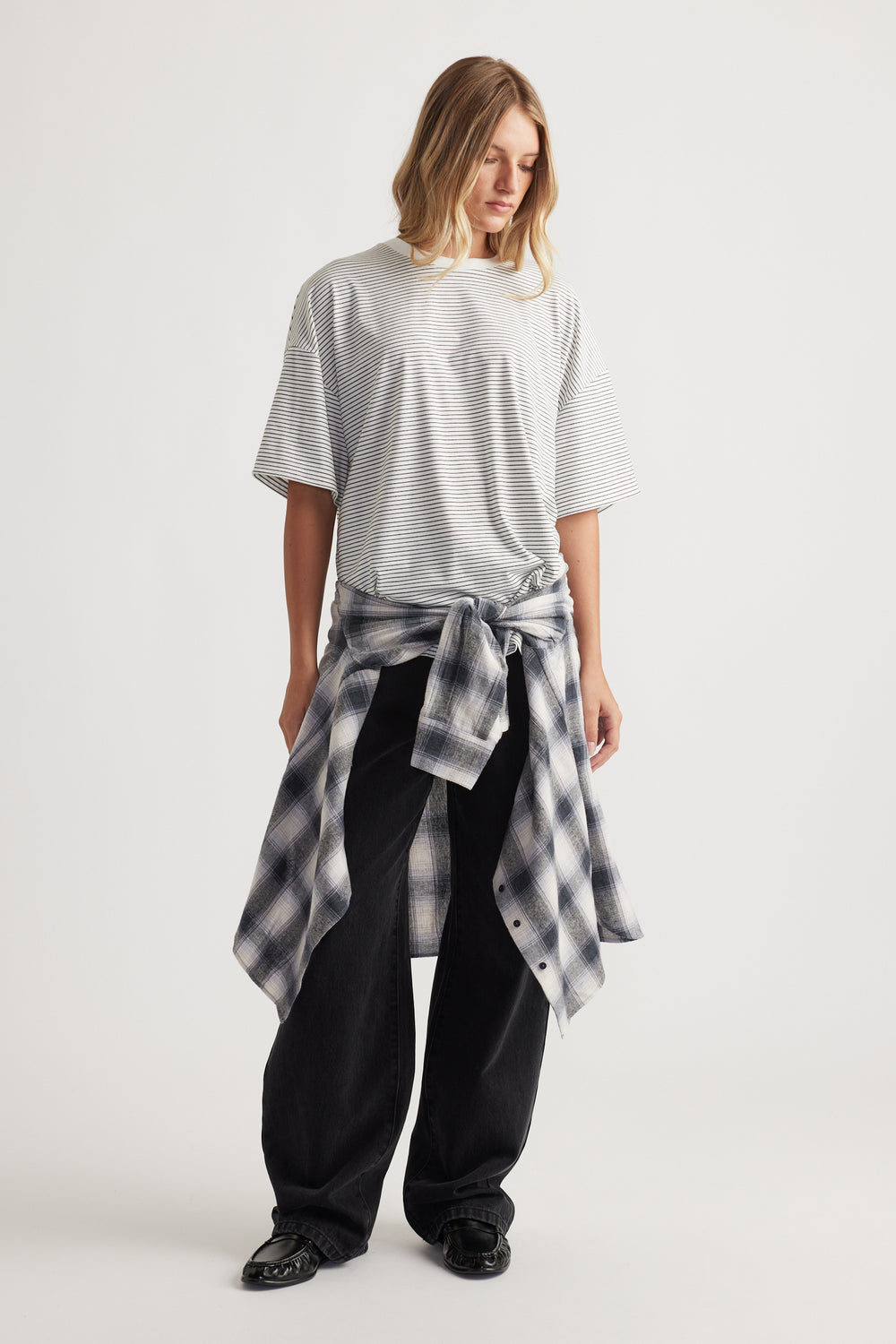Woman wearing a loose striped grey t-shirt with a tied black and white plaid shirt around the waist and black pants
