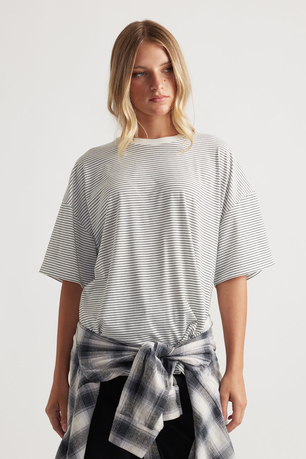 Woman wearing a loose short-sleeve striped top with a plaid shirt tied around the waist against a plain background