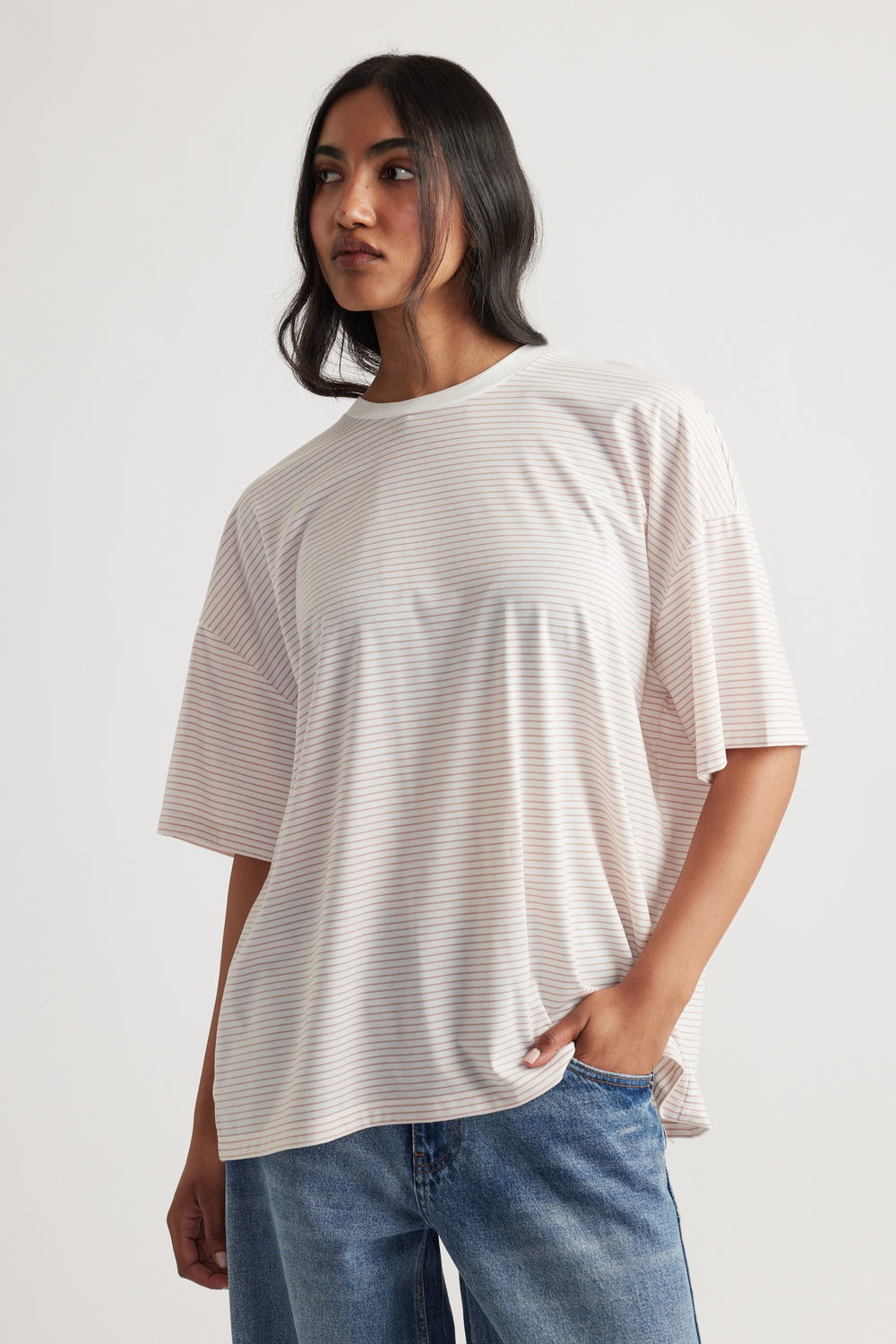 Woman wearing a relaxed fit, light striped T-shirt with half sleeves paired with blue denim jeans, posing against a plain background.