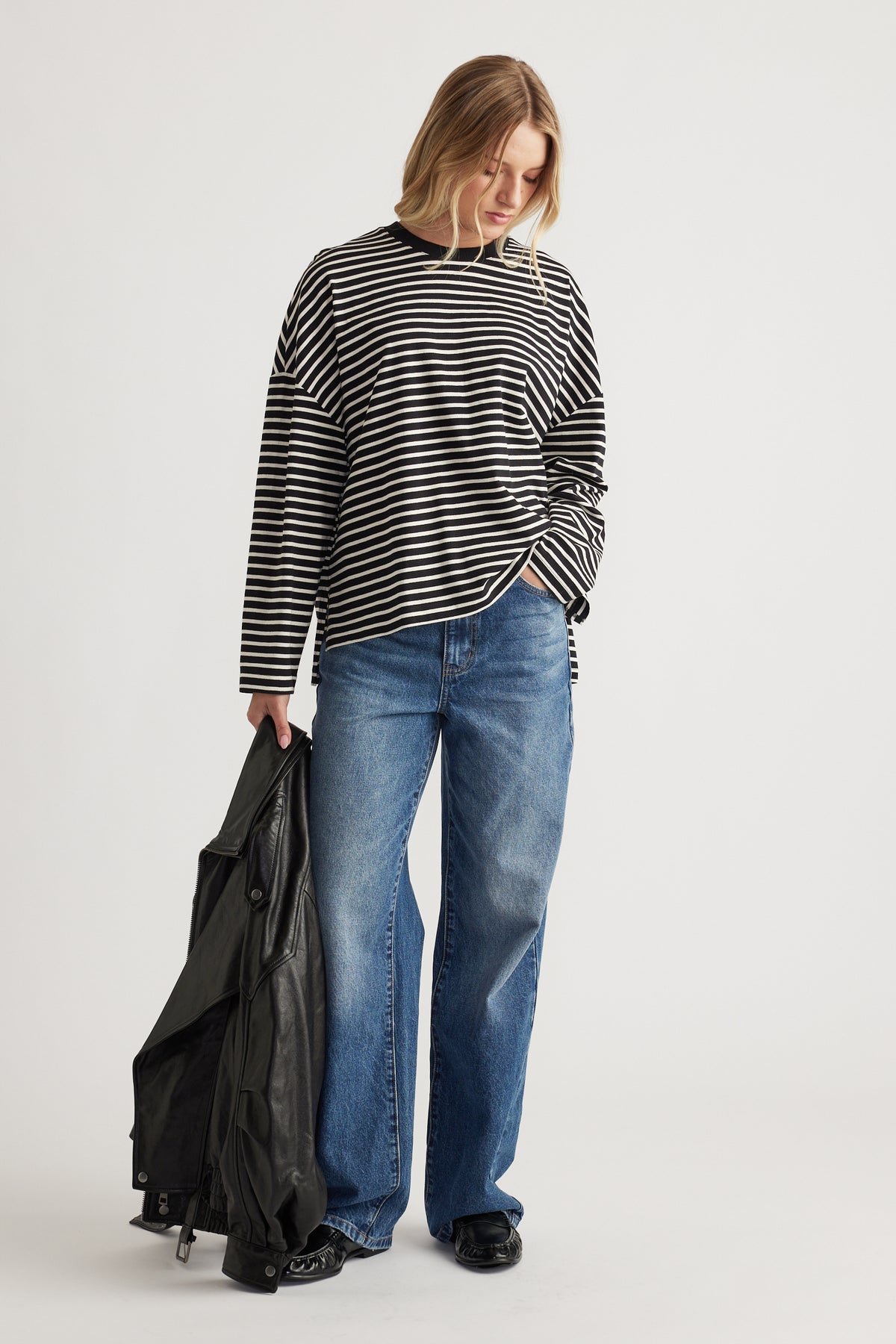 Woman wearing a relaxed black and ivory striped long sleeve top paired with classic blue wide-leg jeans and holding a leather jacket