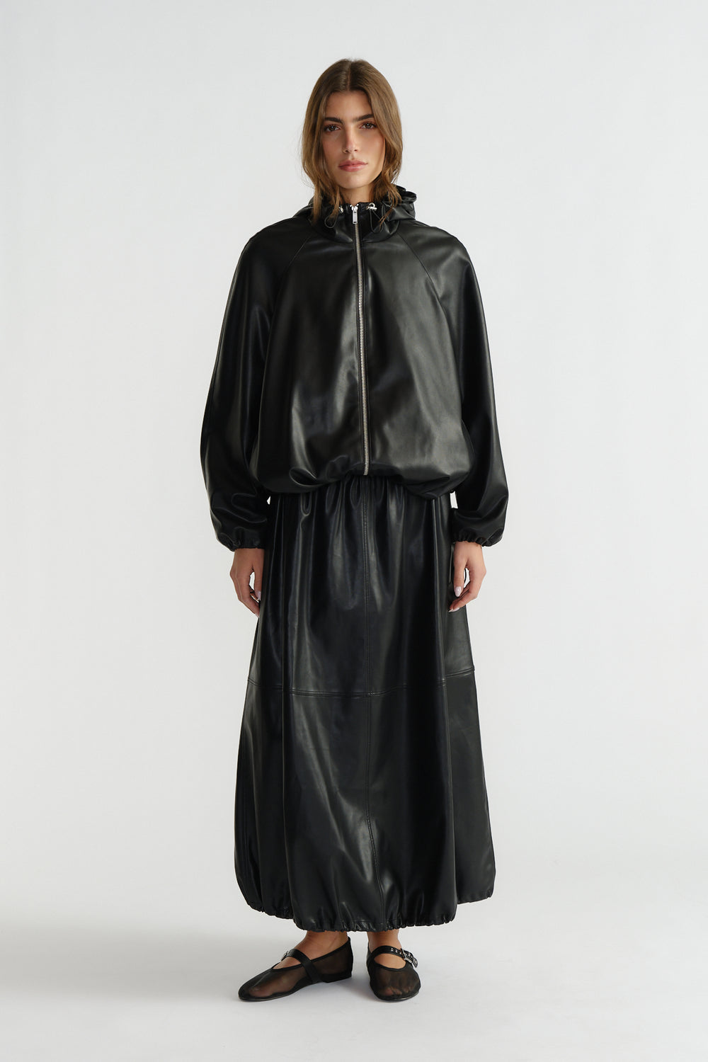 Woman wearing a black faux leather bomber jacket with a matching long skirt and black ballet flats, standing against white.
