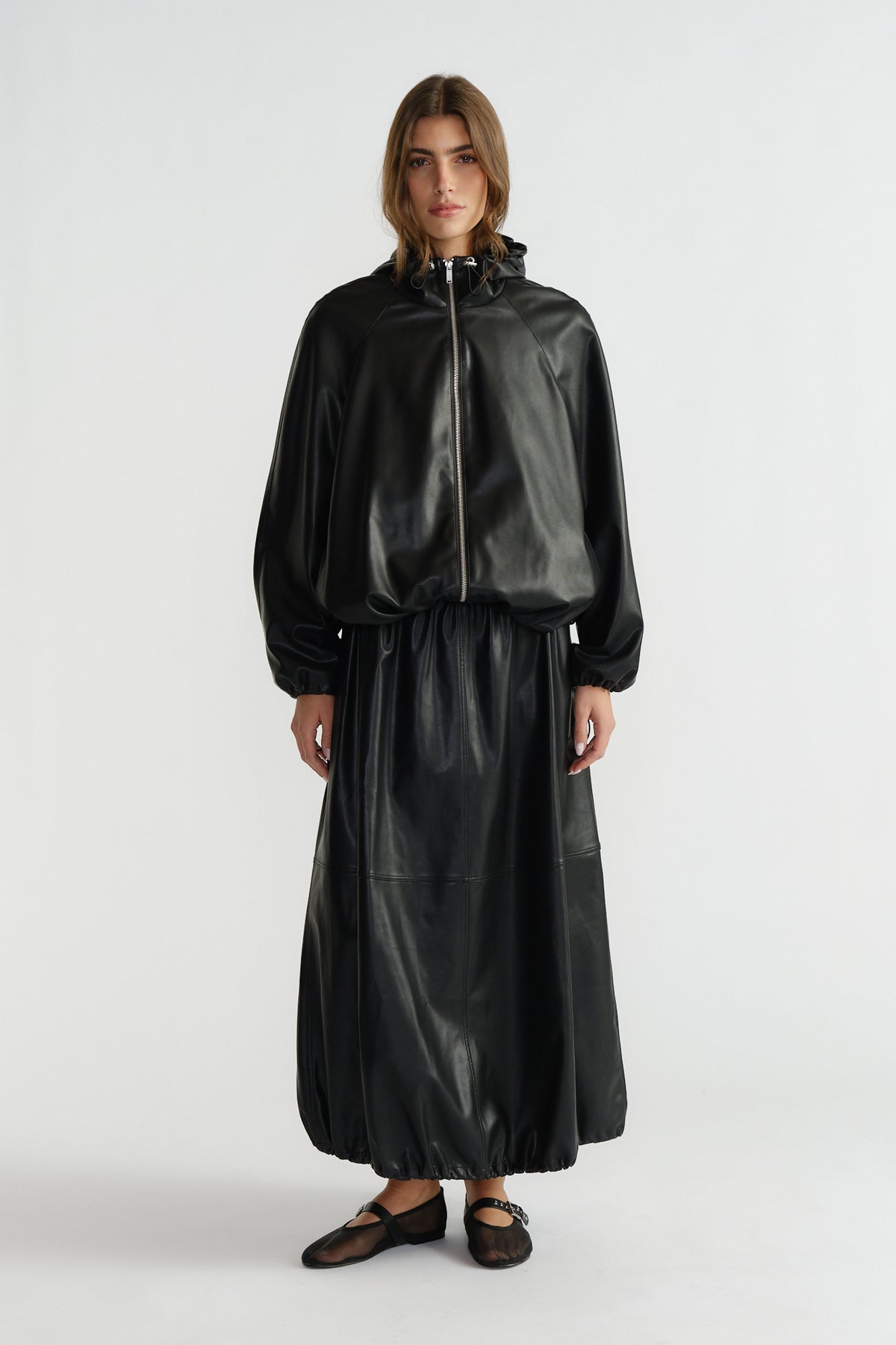Woman wearing a black faux leather bomber jacket with a matching long skirt and black ballet flats, standing against white.