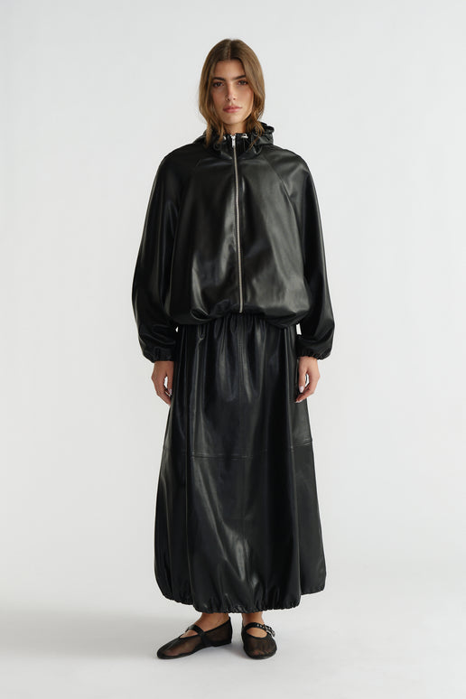 Woman wearing a black faux leather bomber jacket with a matching long skirt and black ballet flats, standing against white.