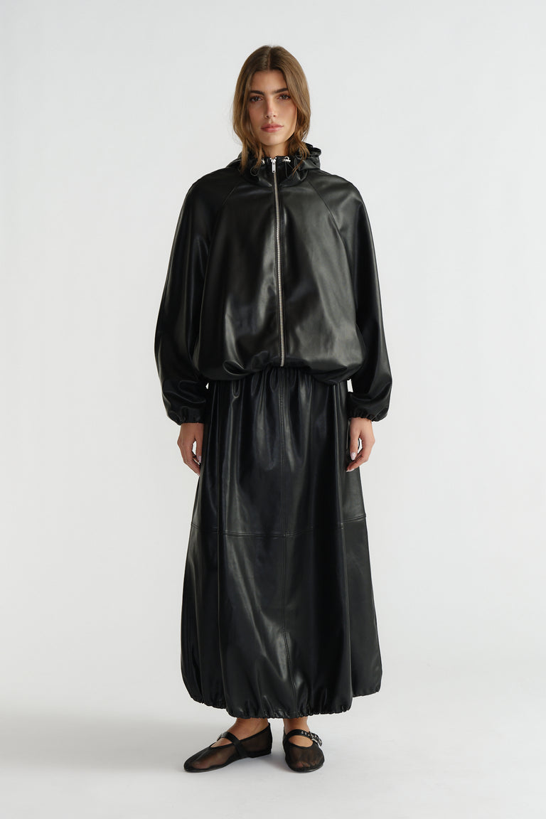 Woman wearing a black faux leather bomber jacket with a matching long skirt and black ballet flats, standing against white.