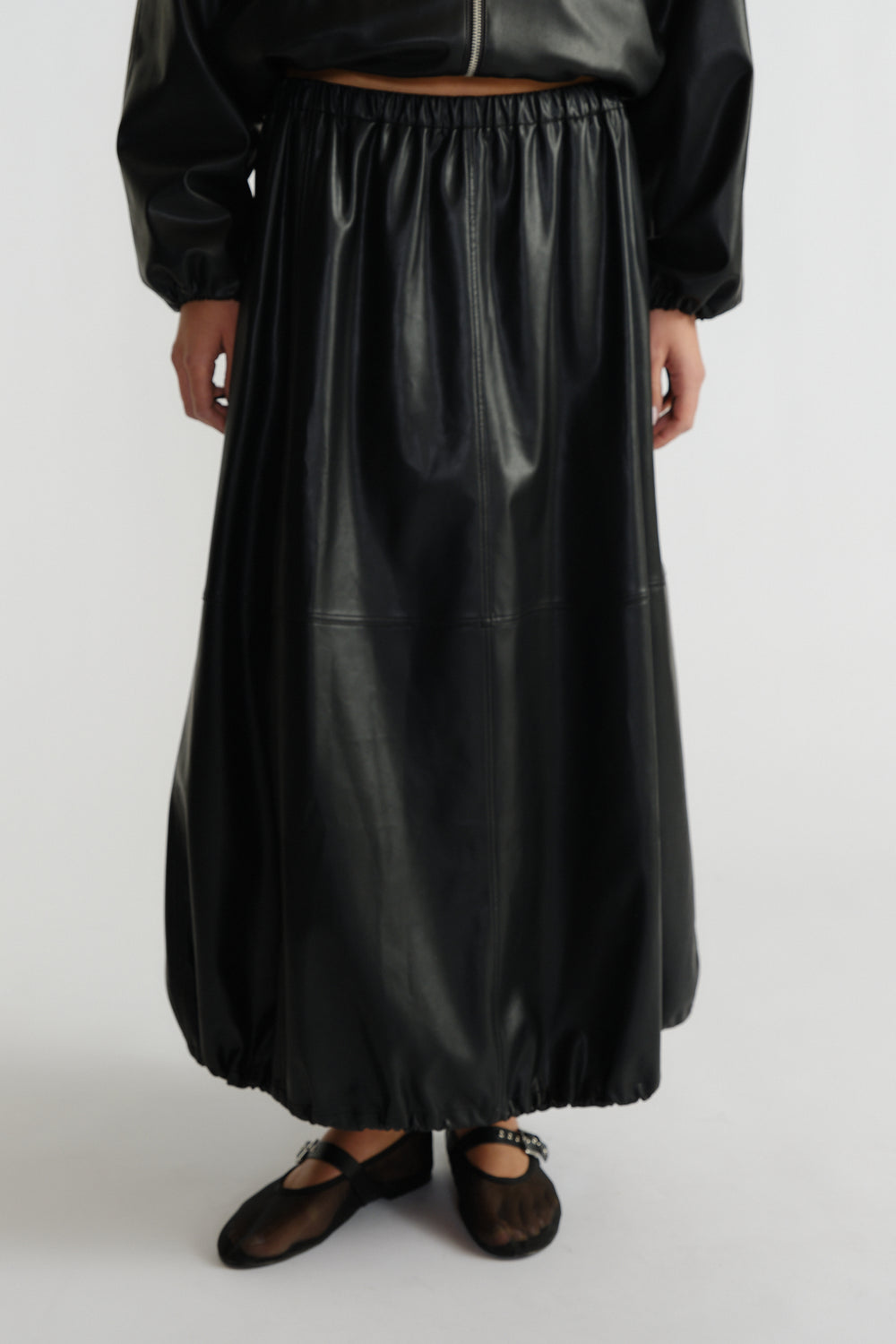 Woman wearing a black faux leather midi skirt with elastic hems paired with sheer black ballet flats and a cropped jacket