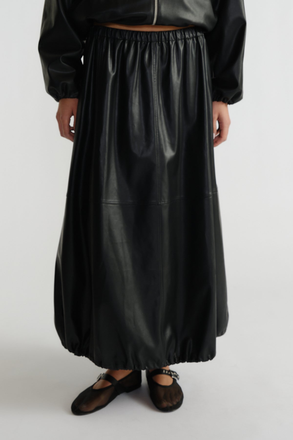 Woman wearing a black faux leather midi skirt with elastic hems paired with sheer black ballet flats and a cropped jacket