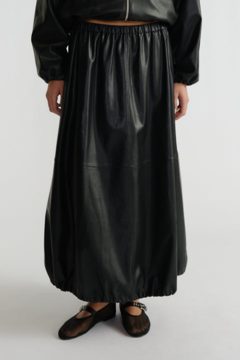 Woman wearing a black faux leather midi skirt with elastic hems paired with sheer black ballet flats and a cropped jacket