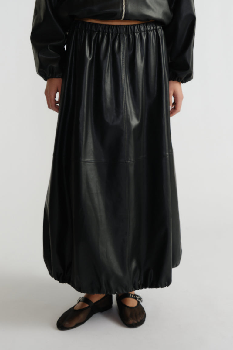 Woman wearing a black faux leather midi skirt with elastic hems paired with sheer black ballet flats and a cropped jacket