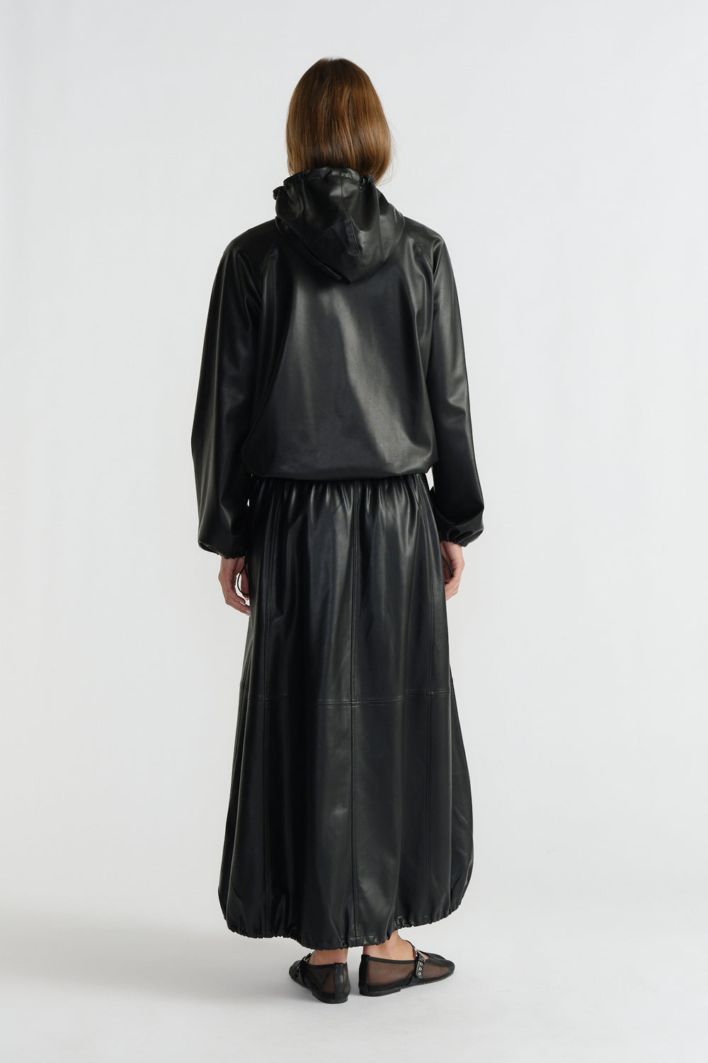 Woman wearing a black faux leather maxi skirt with a gathered hem paired with a relaxed-fit hooded jacket from behind