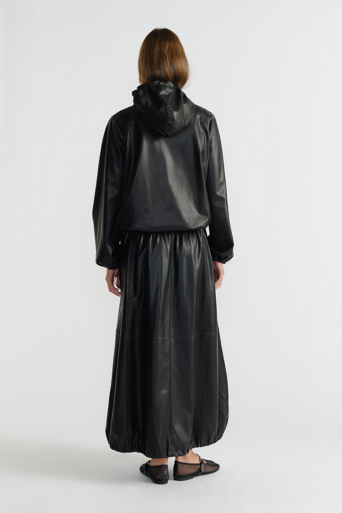 Woman wearing a black faux leather maxi skirt with a gathered hem paired with a relaxed-fit hooded jacket from behind