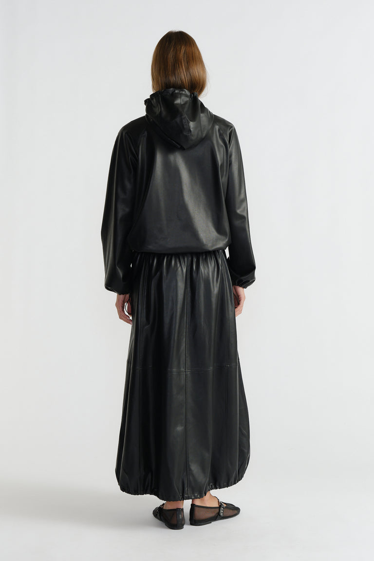Woman wearing a black faux leather maxi skirt with a gathered hem paired with a relaxed-fit hooded jacket from behind