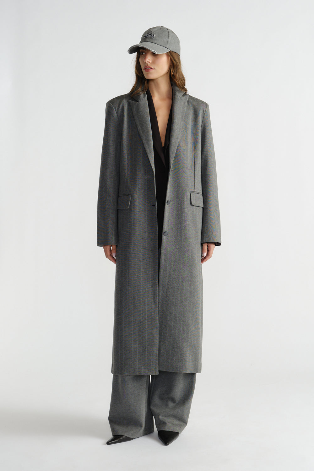 Woman wearing a long charcoal pinstripe blazer coat with matching wide-leg pants and a grey baseball cap.