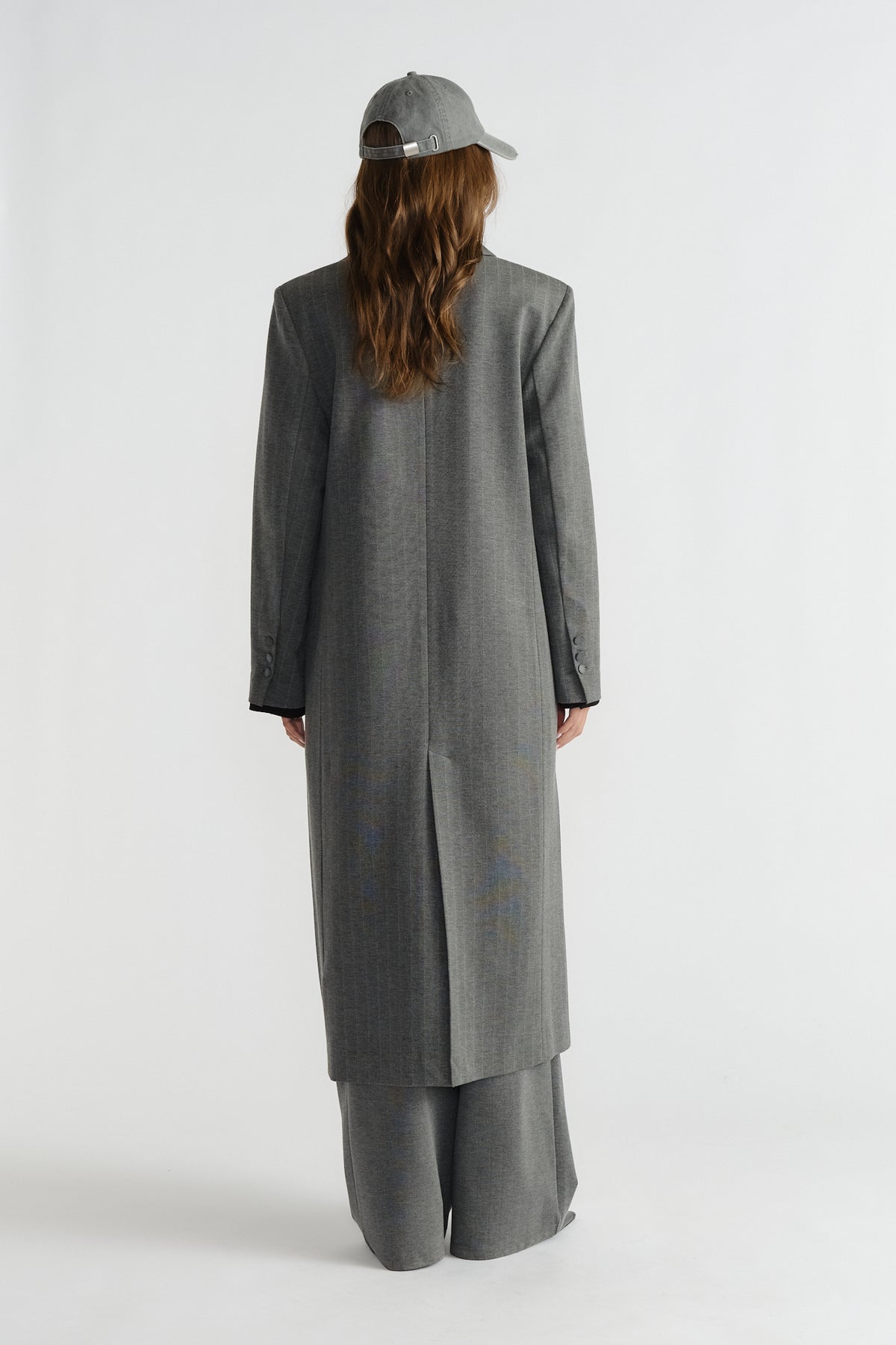 Back view of a long grey pinstripe blazer coat paired with matching wide-leg pants and a casual grey cap.