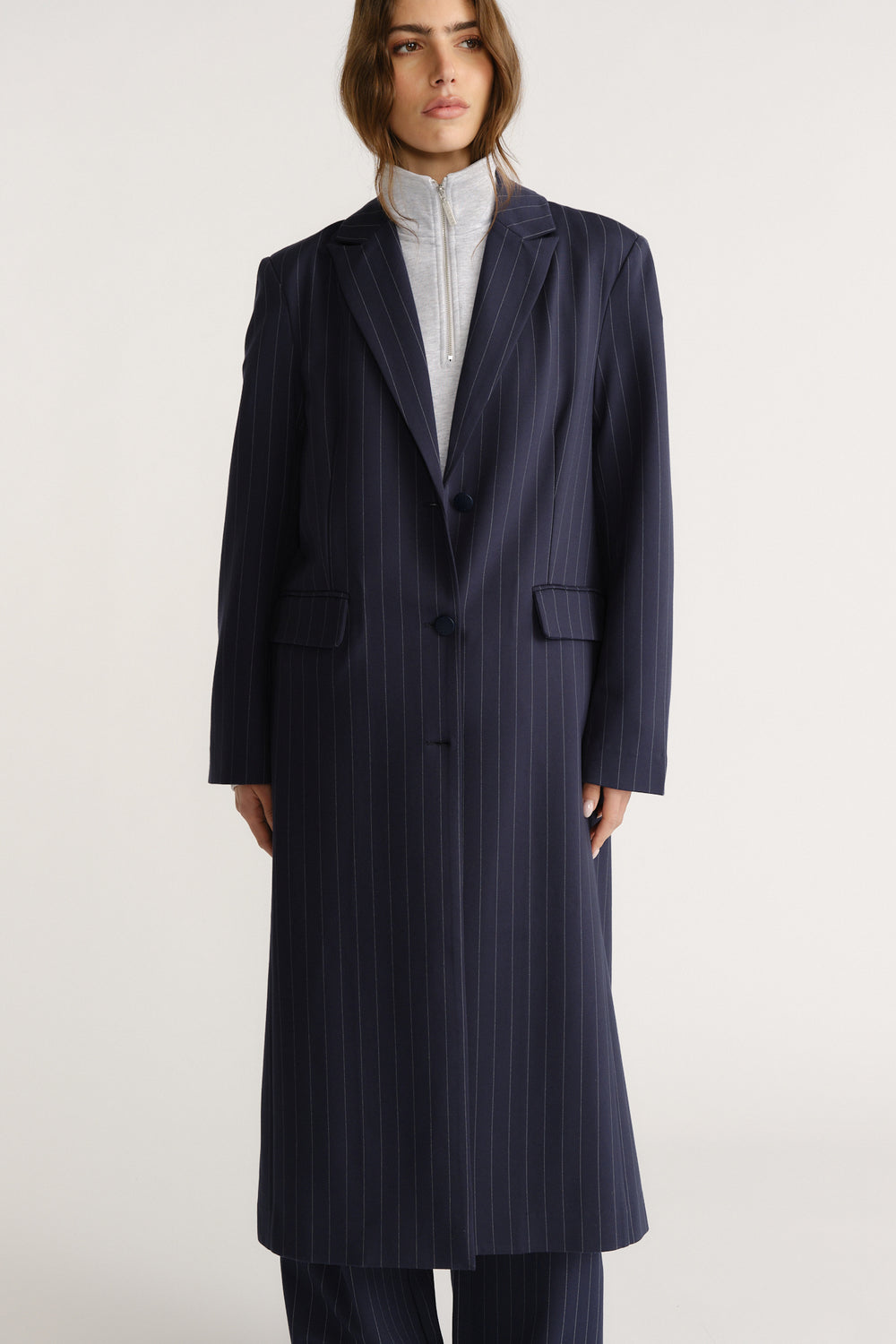 Woman wearing a tailored navy pinstripe long blazer coat layered over a light zip-up top for a polished look