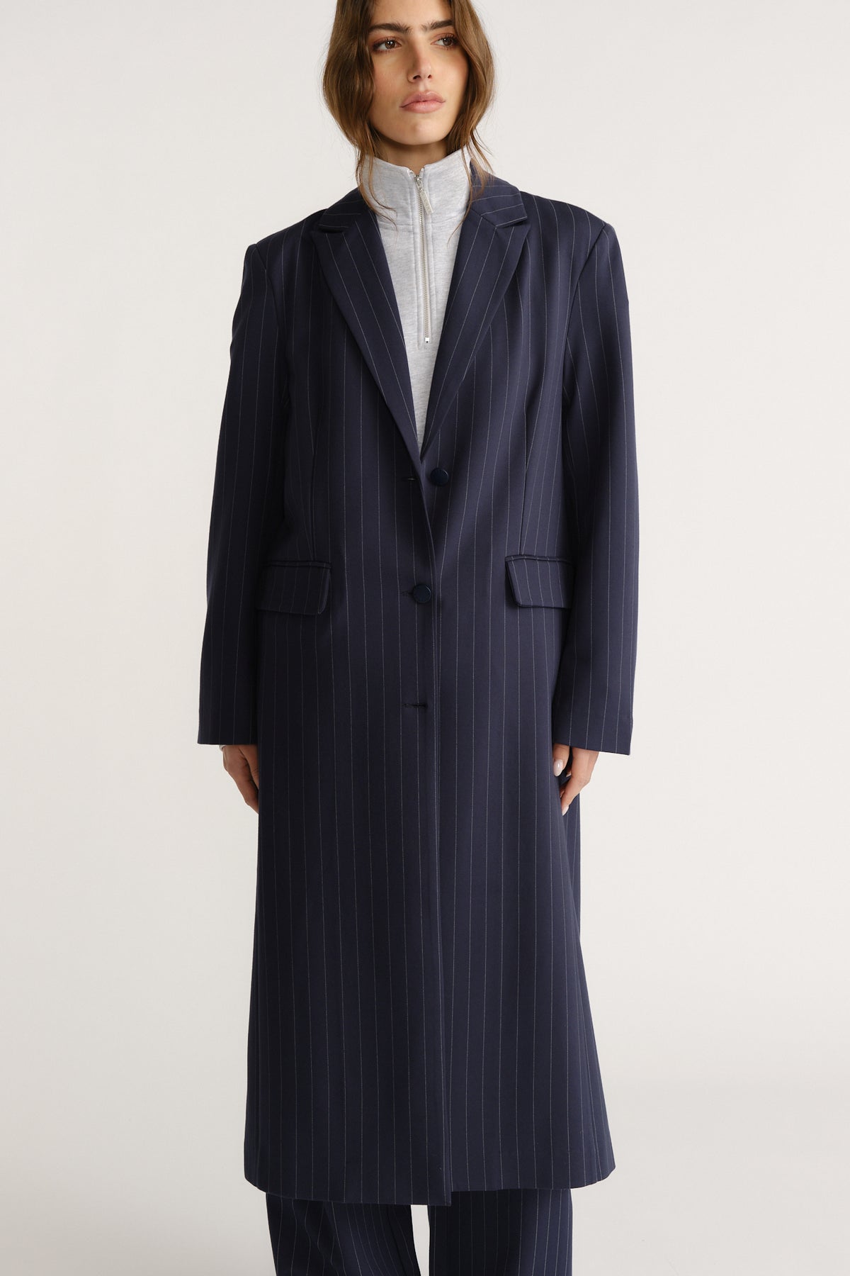 Woman wearing a tailored navy pinstripe long blazer coat layered over a light zip-up top for a polished look