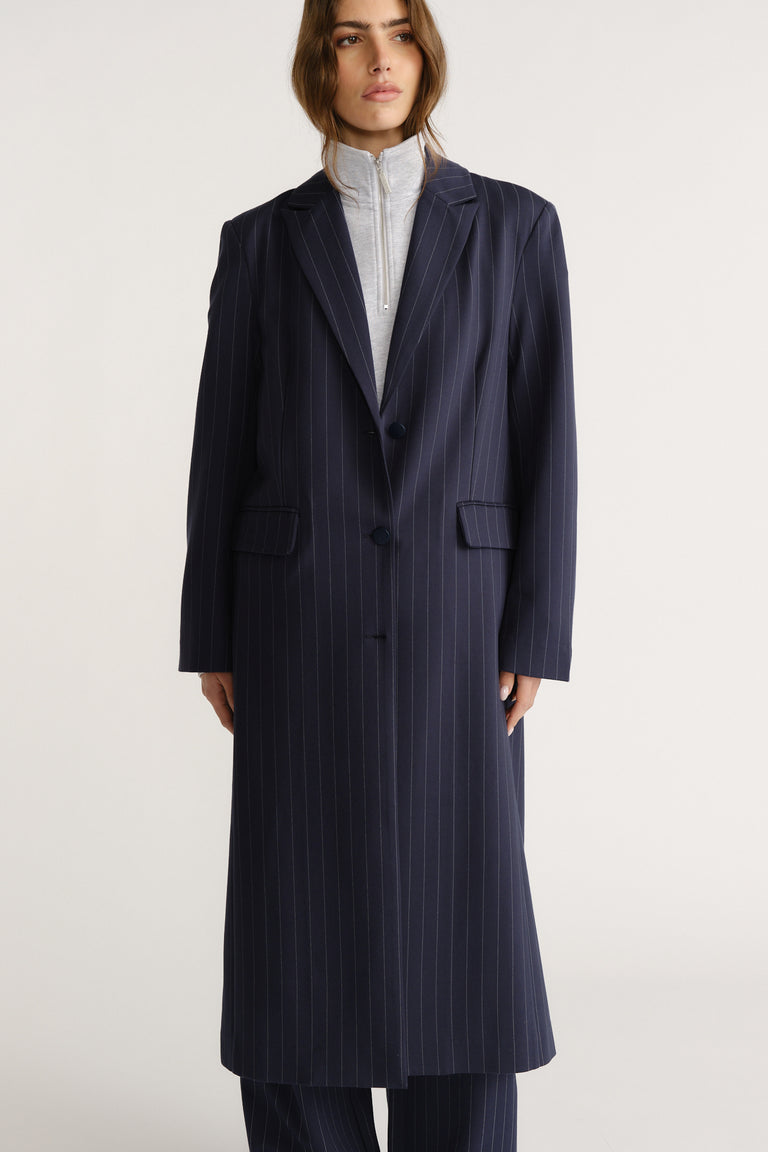 Woman wearing a tailored navy pinstripe long blazer coat layered over a light zip-up top for a polished look
