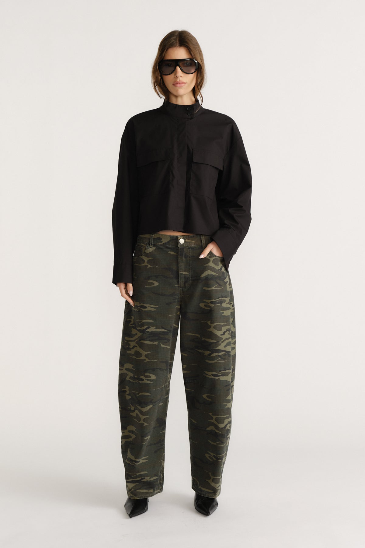 Woman wearing a cropped black utility shirt styled with loose-fitting camo pants and black pointed boots, accessorized with sunglasses.
