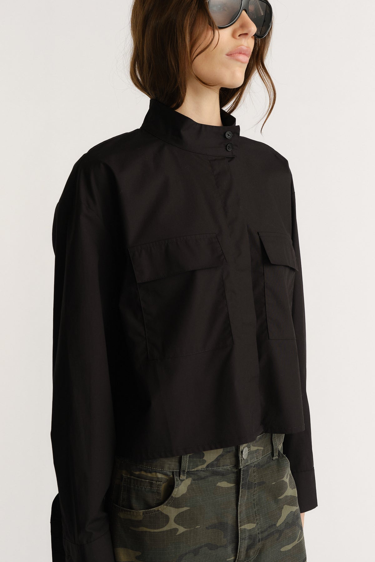 Woman wearing a black utility-style shirt with a high collar and oversized pockets paired with camo pants and sunglasses