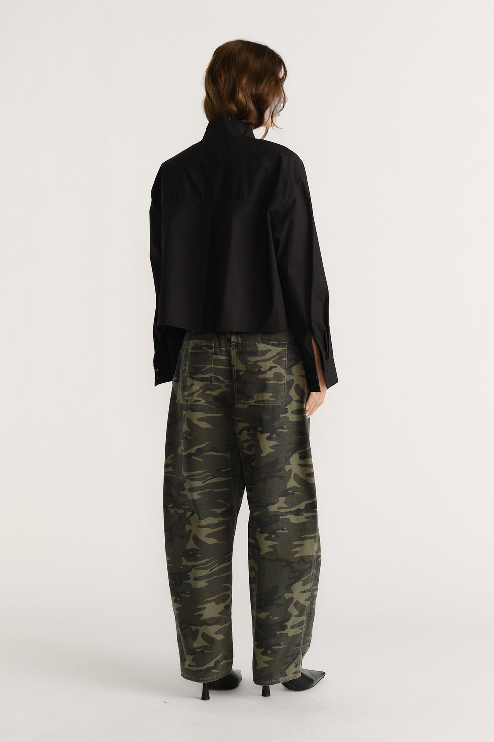 Woman seen from behind wearing a black oversized shirt and loose camouflage pants with black heeled boots.