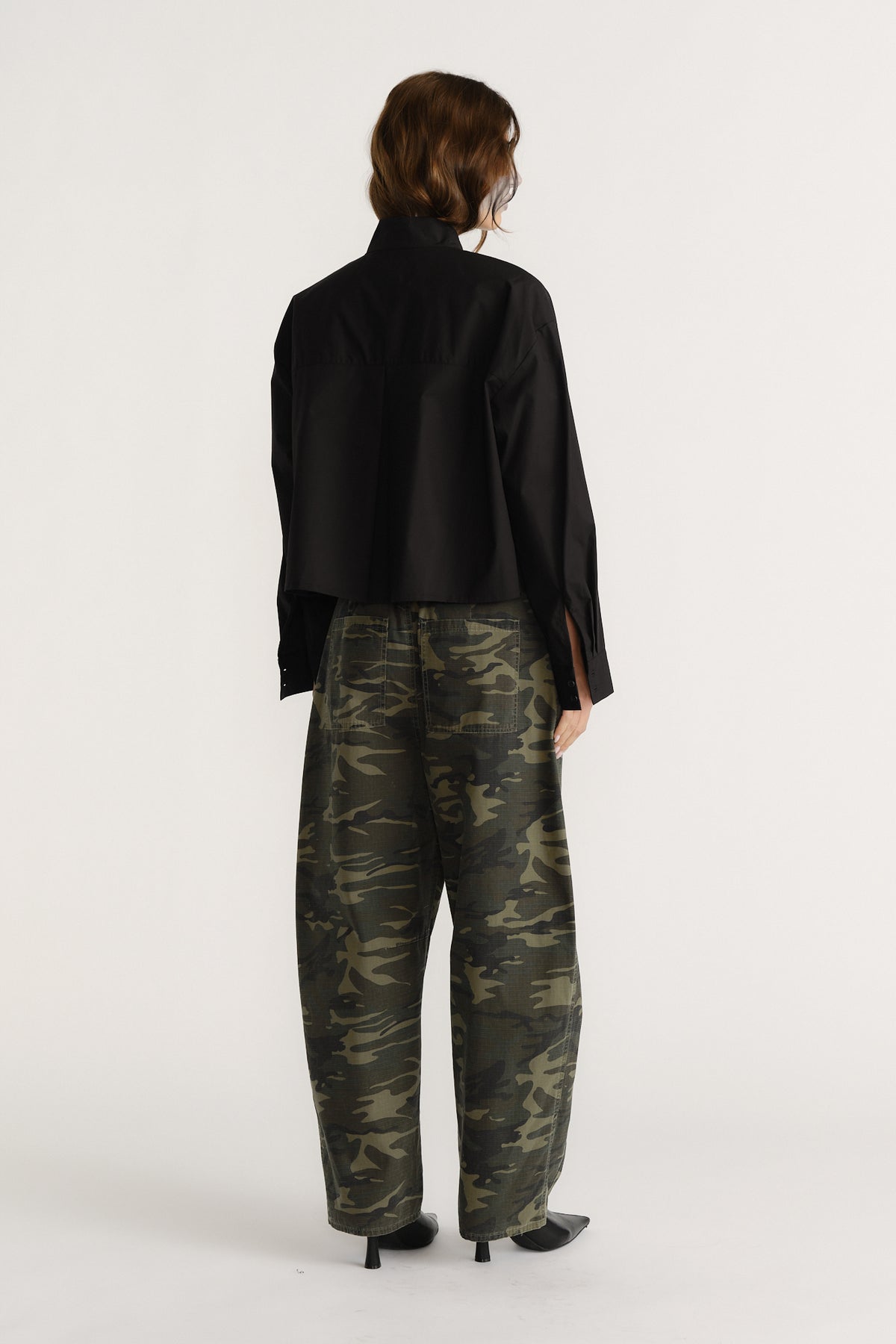 Woman seen from behind wearing a black oversized shirt and loose camouflage pants with black heeled boots.