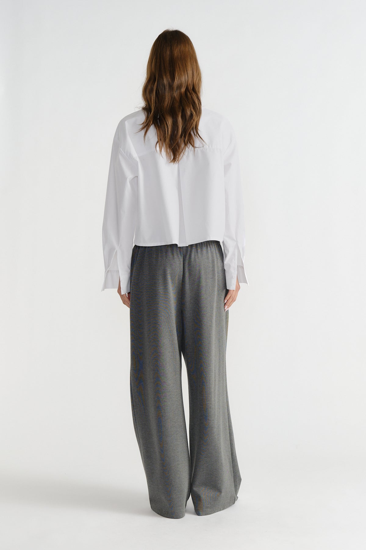 Back view of a woman wearing a relaxed white shirt paired with loose, wide-legged dark grey trousers.