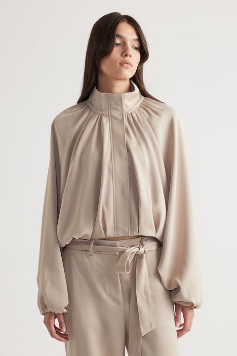 Woman wearing a soft beige satin bubble jacket with gathered detailing and matching high-waisted pants with a tie belt