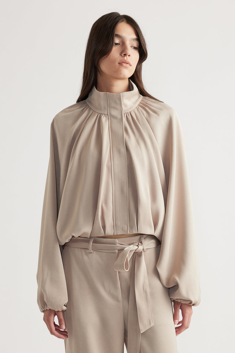 Woman wearing a soft beige satin bubble jacket with gathered detailing and matching high-waisted pants with a tie belt