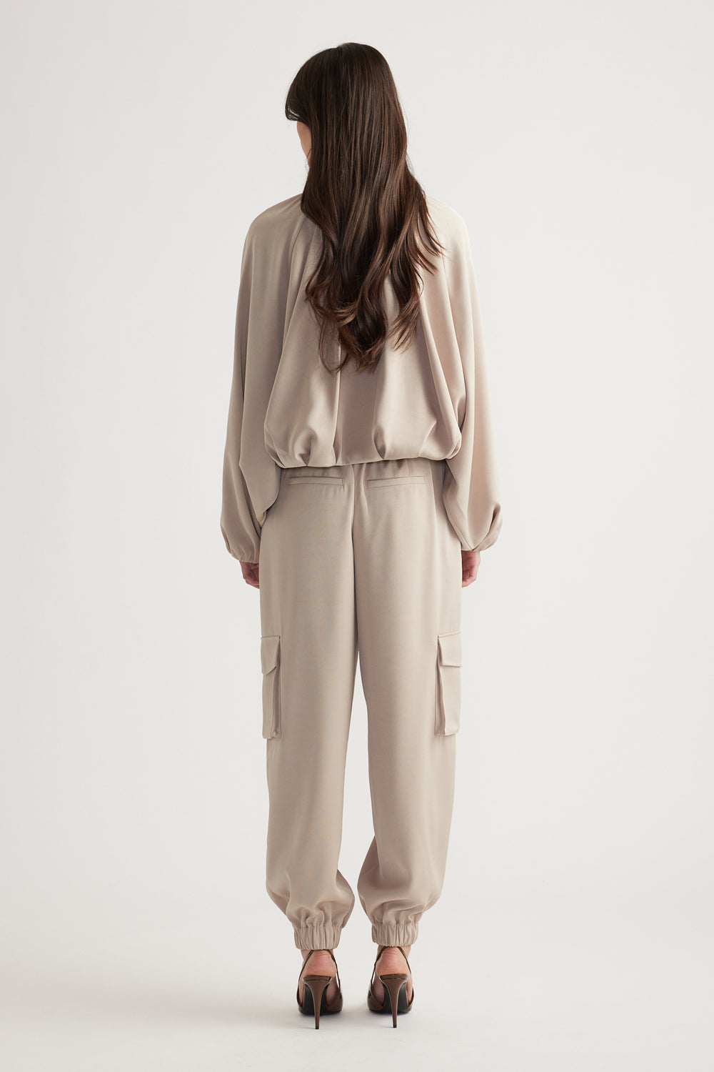 Woman wearing beige relaxed-fit cargo pants and a loose satin bubble jacket paired with black heeled sandals, back view