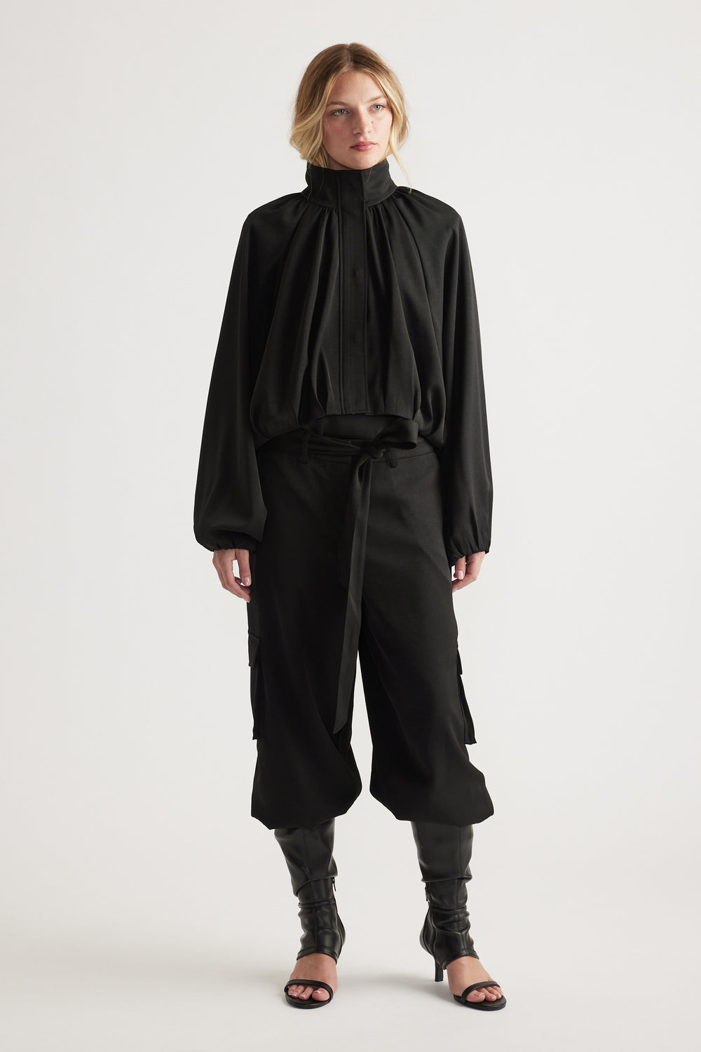 Woman wearing a black high-neck satin jacket paired with wide-leg cropped pants and black heeled sandals
