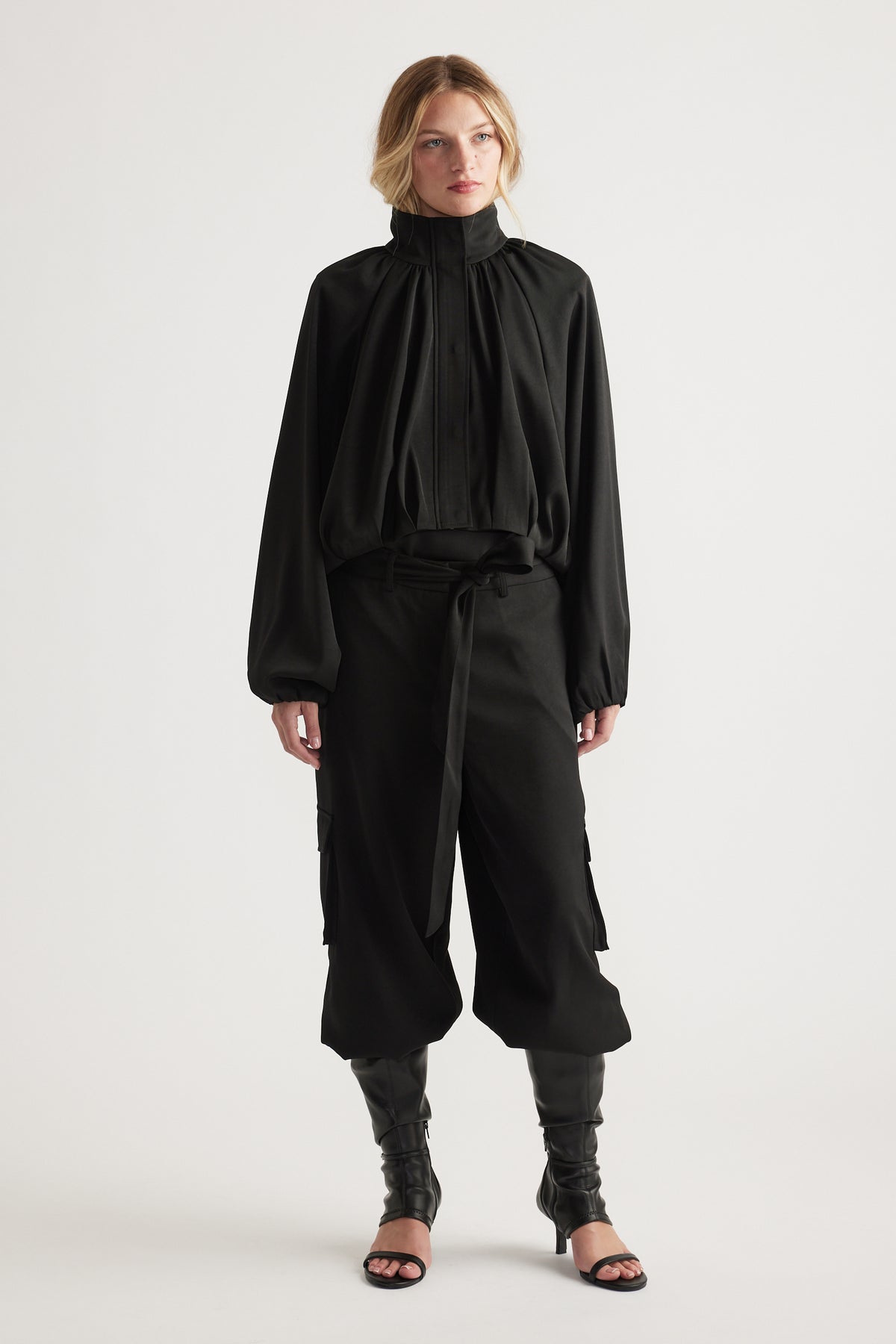 Woman wearing a black high-neck satin jacket paired with wide-leg cropped pants and black heeled sandals