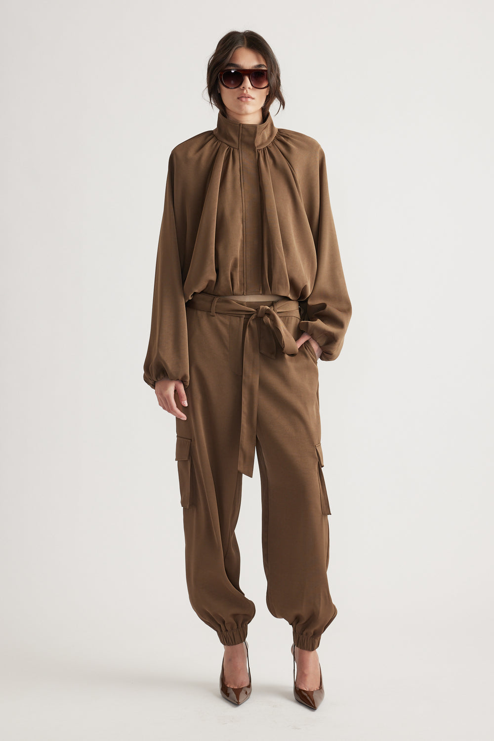 Woman in bronze satin bubble jacket and matching belted cargo pants, styled with sunglasses and heels for a polished look