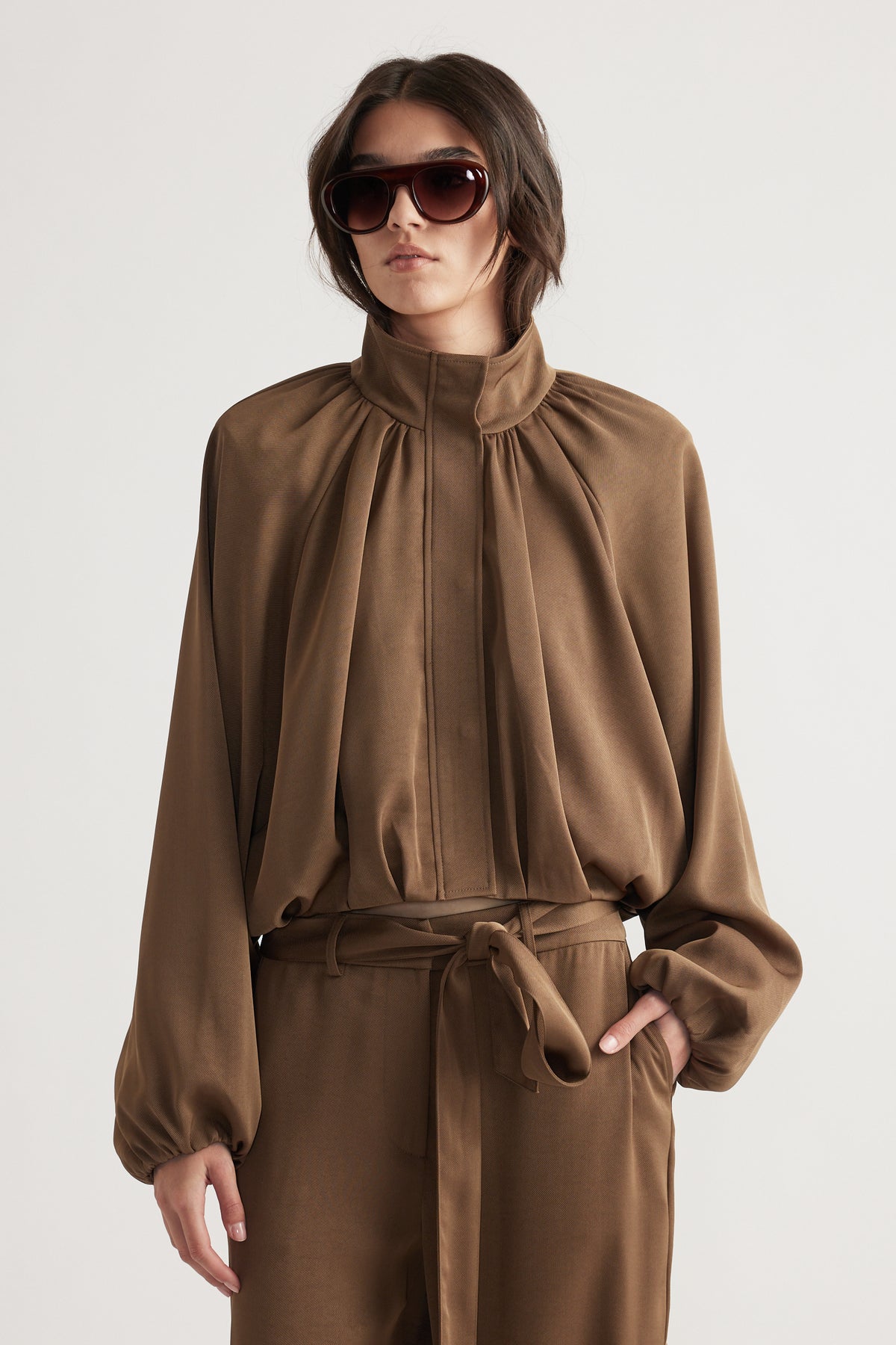 Woman wearing a bronze-toned satin jacket with gathered detailing and a high collar, styled with matching belted pants