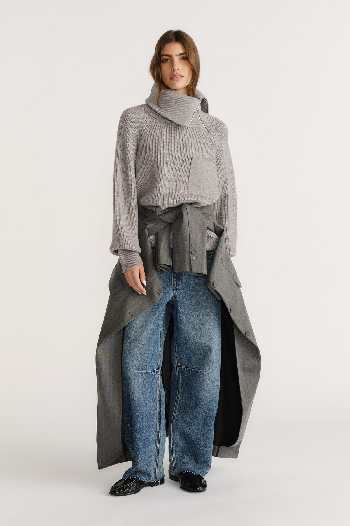 Woman wearing a grey high-neck knit jumper styled with wide-leg denim jeans and a grey coat tied at the waist