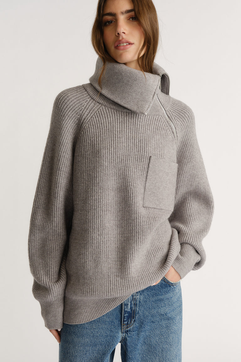 Woman wearing a grey ribbed knit jumper with a high collar and front pocket paired with relaxed blue jeans