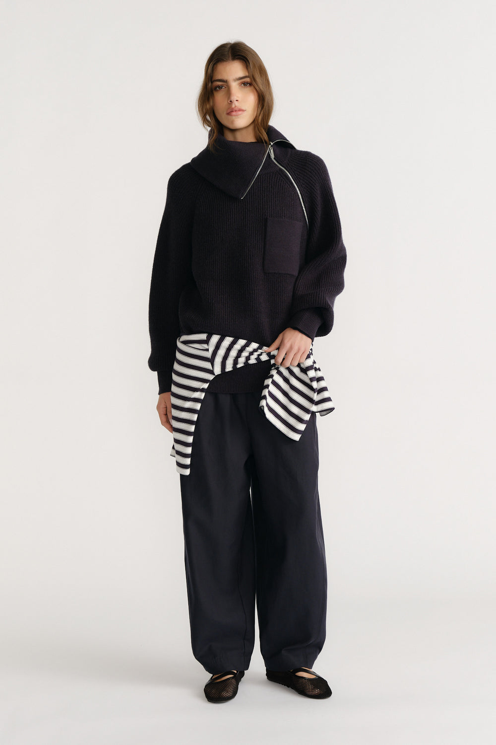 Woman wearing a navy high neck knit jumper with oversized collar, wide-leg pants, and a striped shirt tied at the waist