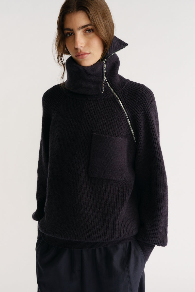 Woman wearing a navy high-neck knit jumper with an asymmetric zip and front patch pocket paired with relaxed trousers