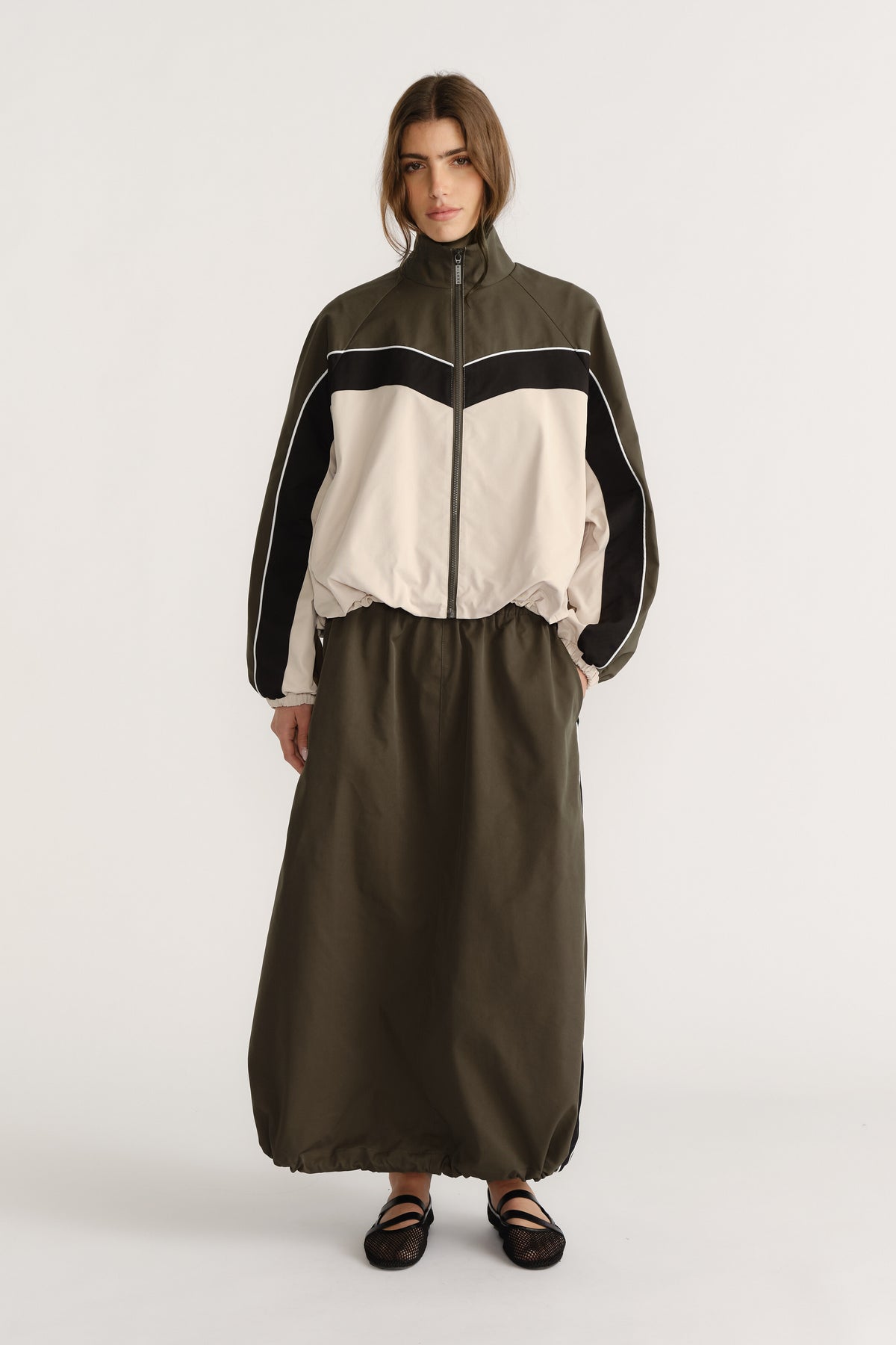 Front view of woman wearing oversized khaki and cream windbreaker with contrast panels and long olive skirt