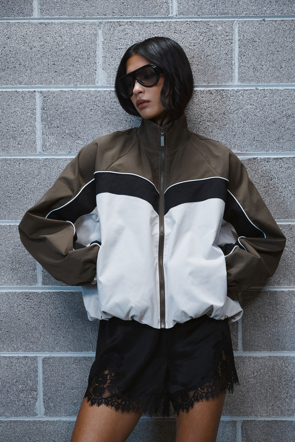 Woman in an oversized brown, black and white windbreaker with black lace trimmed shorts and sunglasses