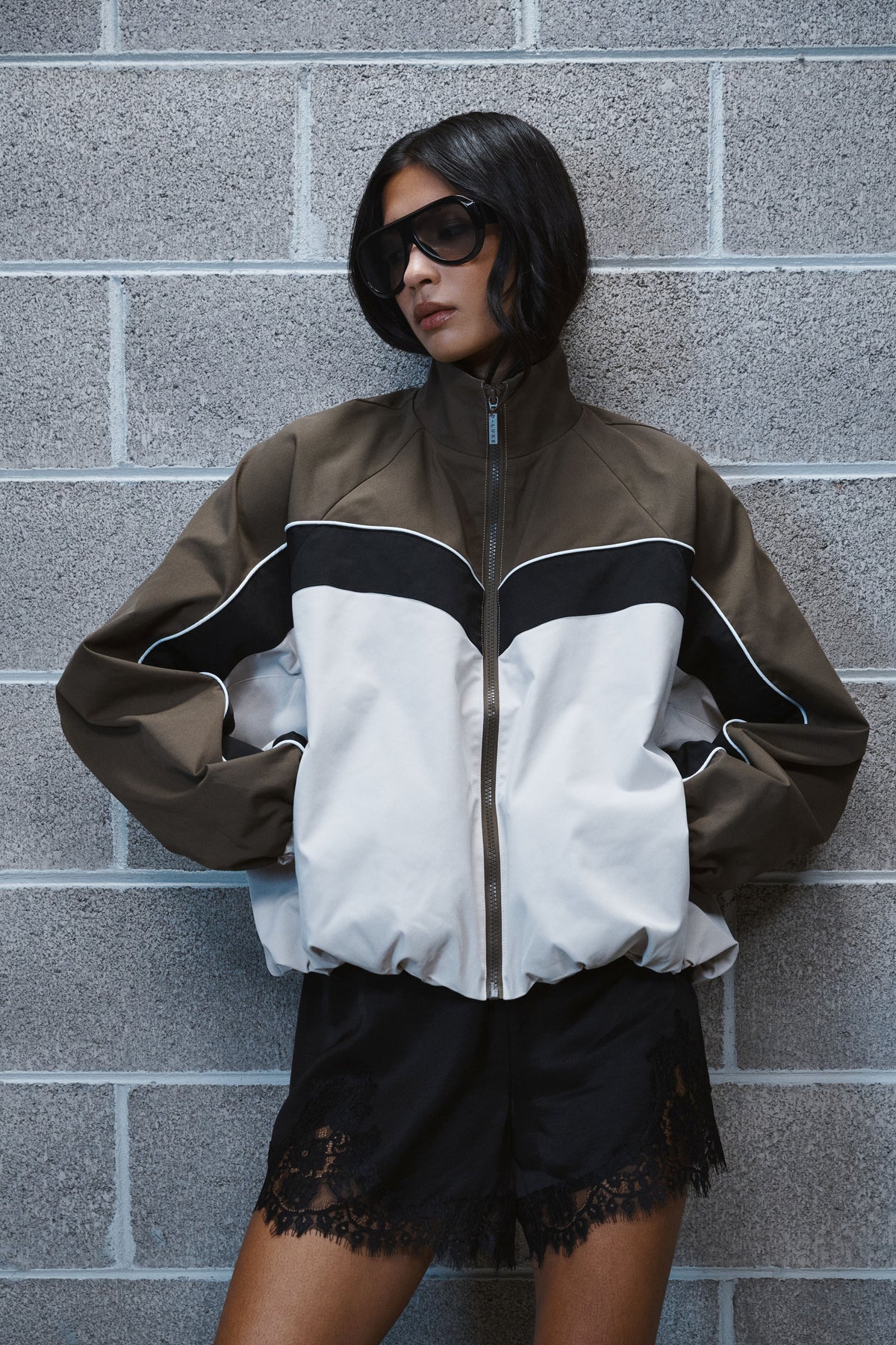 Woman in an oversized brown, black and white windbreaker with black lace trimmed shorts and sunglasses