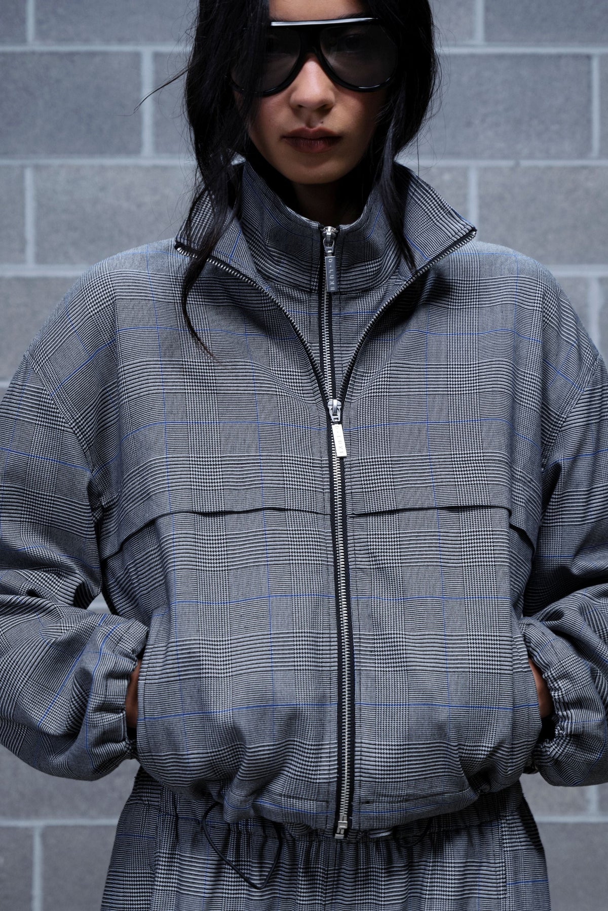Woman wearing a grey check zip up jacket with a high neck, front pockets and adjustable hem