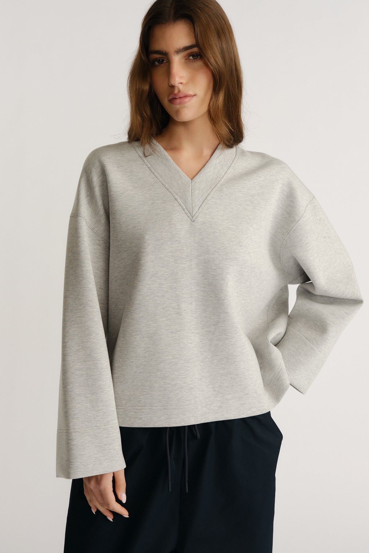 Woman wearing a relaxed light grey V-neck neoprene top with drop shoulders and wide sleeves paired with dark pants