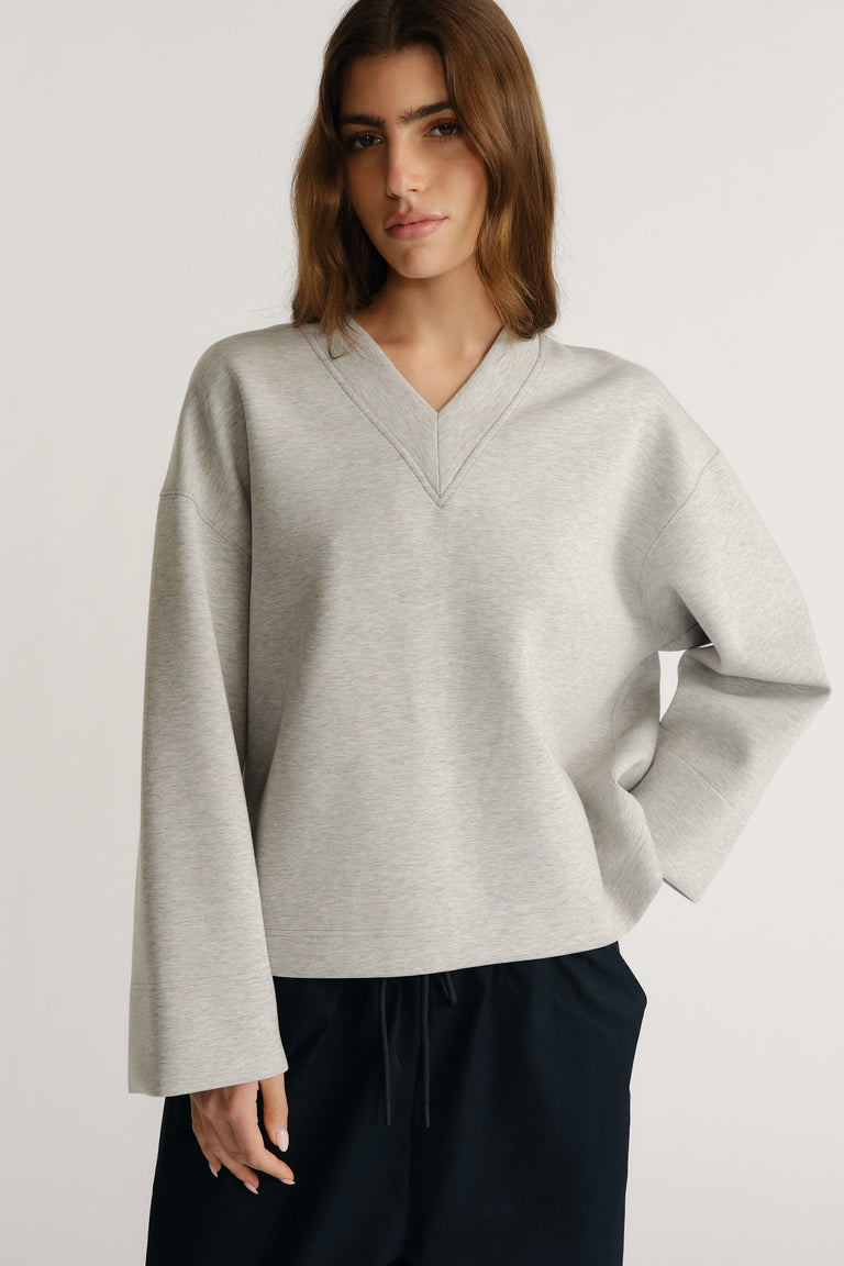 Woman wearing a relaxed light grey V-neck neoprene top with drop shoulders and wide sleeves paired with dark pants