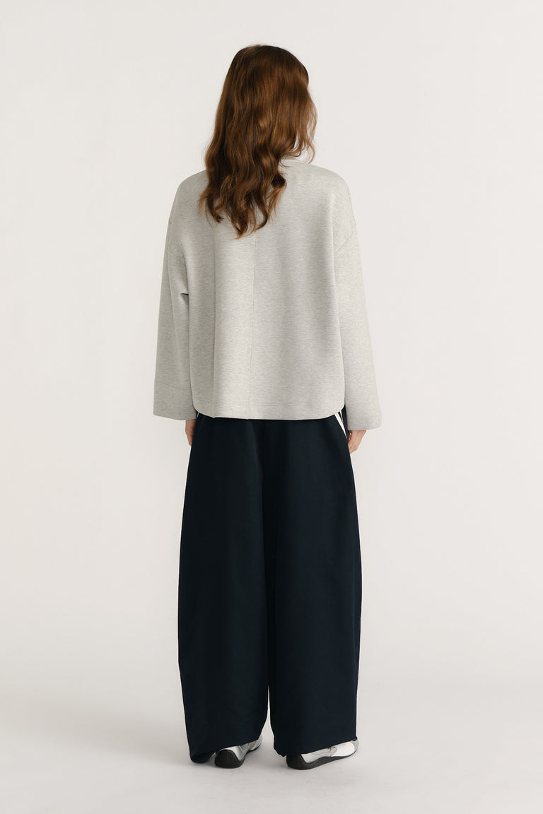 Woman wearing relaxed light grey neoprene V-neck sweat paired with loose wide-leg black pants viewed from behind