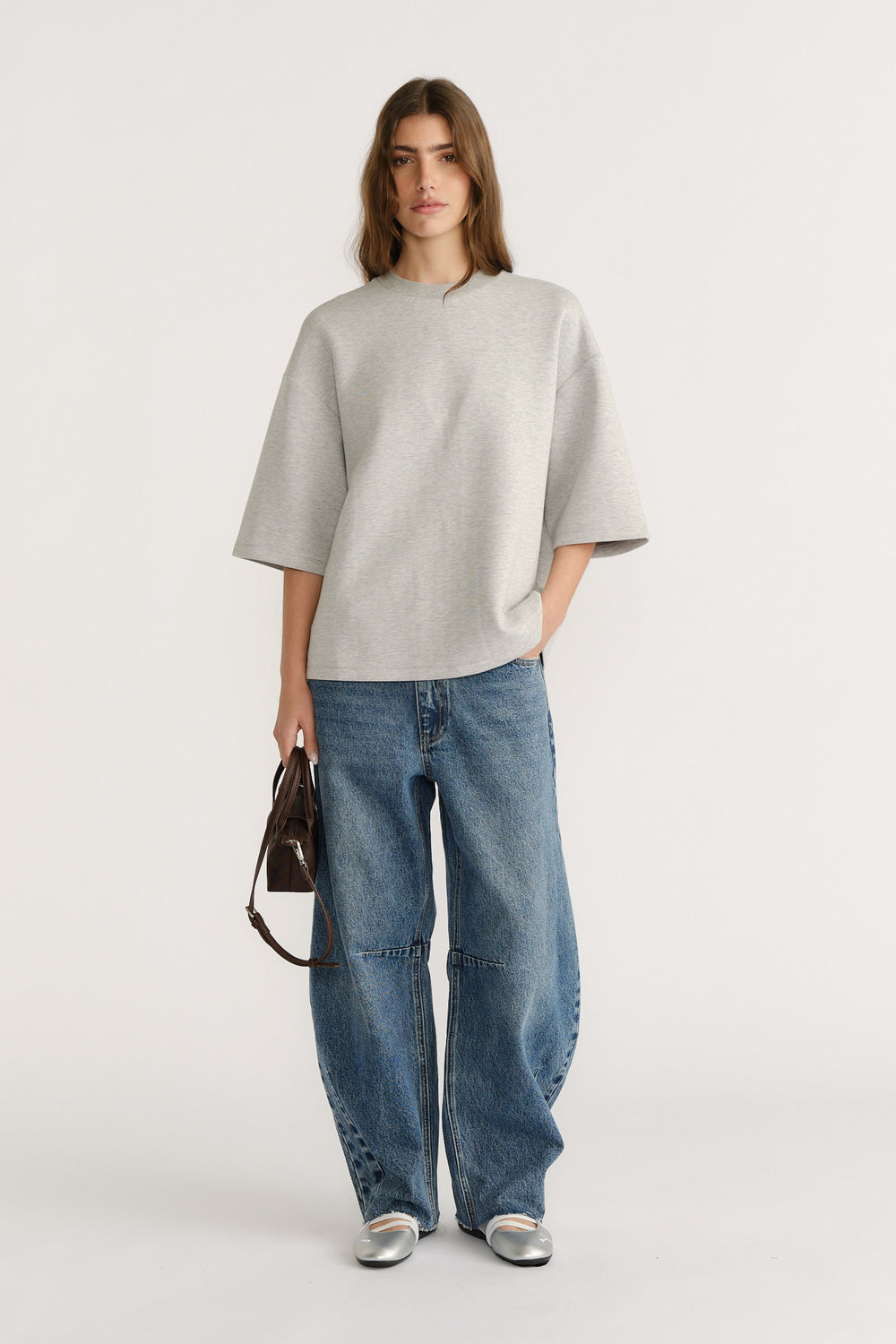 Woman wearing oversized light grey neoprene tee with drop shoulders paired with relaxed wide-leg blue jeans and silver flats
