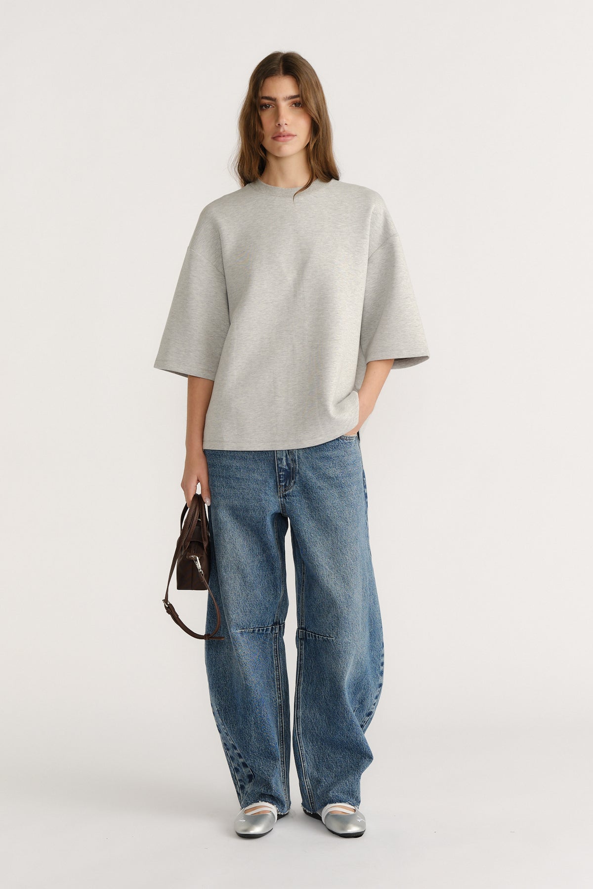 Woman wearing oversized light grey neoprene tee with drop shoulders paired with relaxed wide-leg blue jeans and silver flats