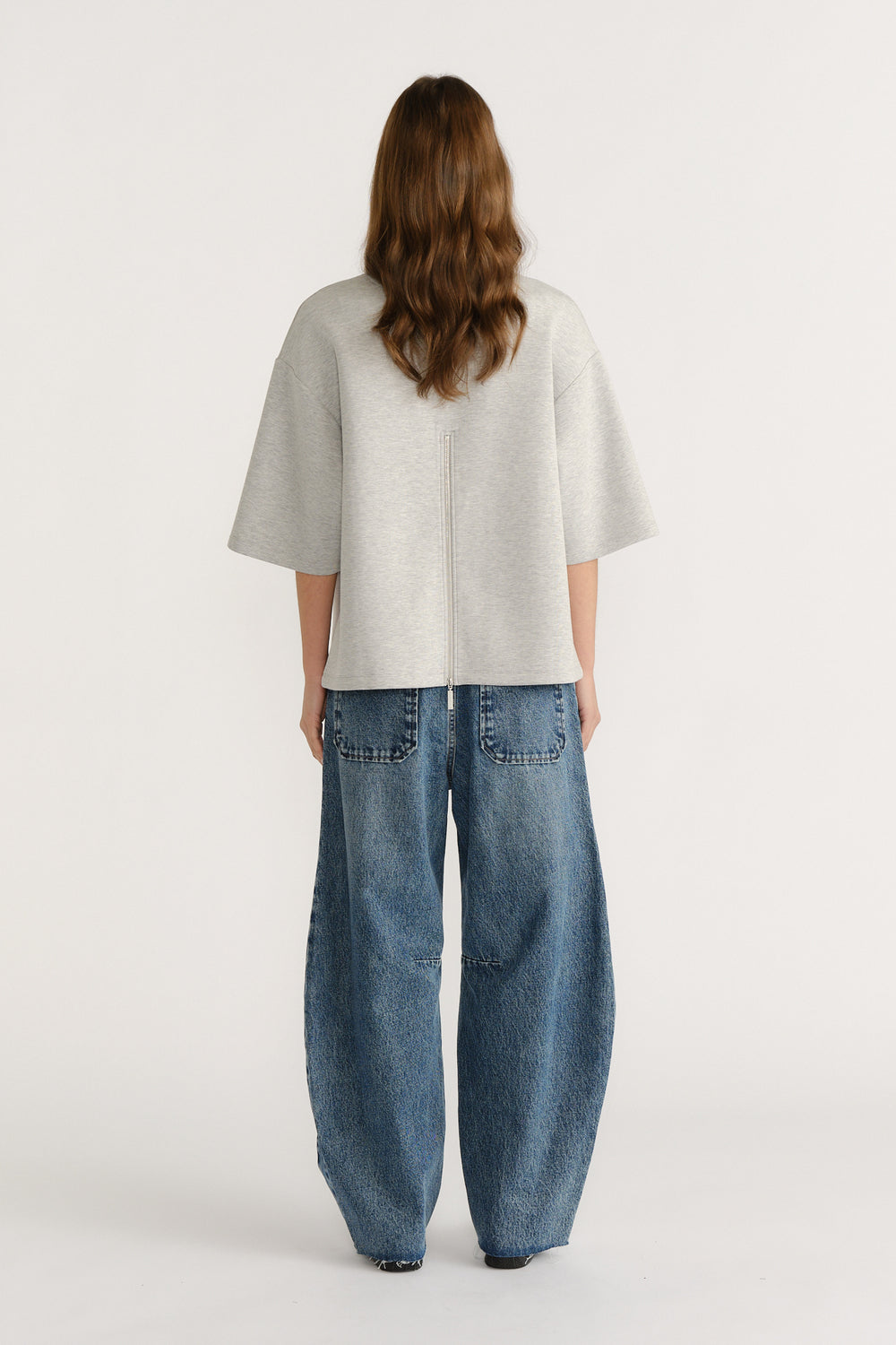 Woman wearing oversized light grey short sleeve top with back zip detail paired with loose wide-leg blue jeans